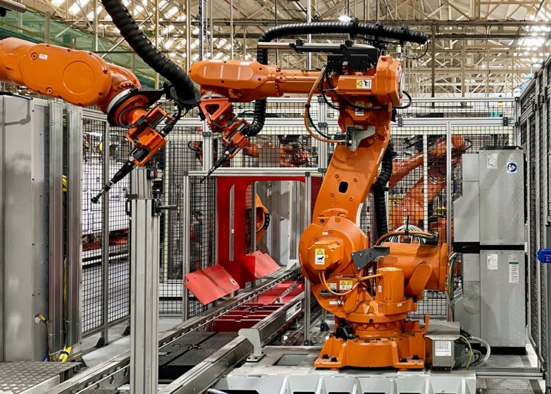 Orange industrial robots operate inside a factory, surrounded by metal cages and equipment. They are part of an automated assembly line.