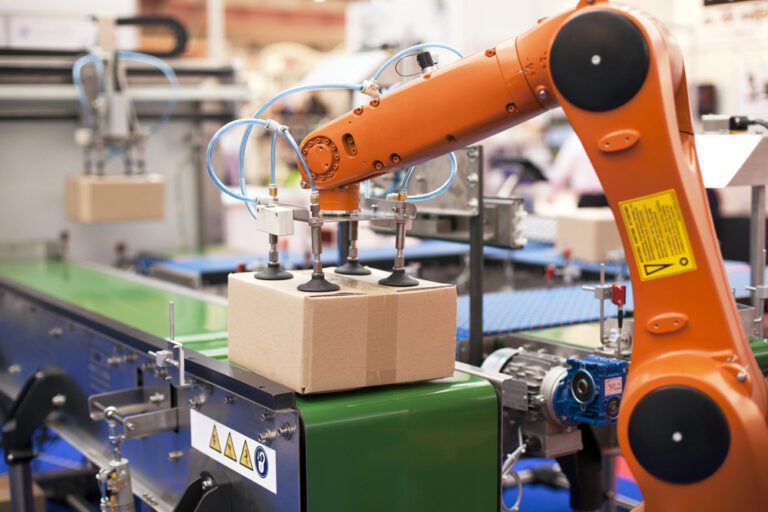 An orange robotic arm is seen placing a cardboard box onto a conveyor belt in an industrial setting, seamlessly blending efficiency that wouldn't look out of place even in the most advanced home automation setups.