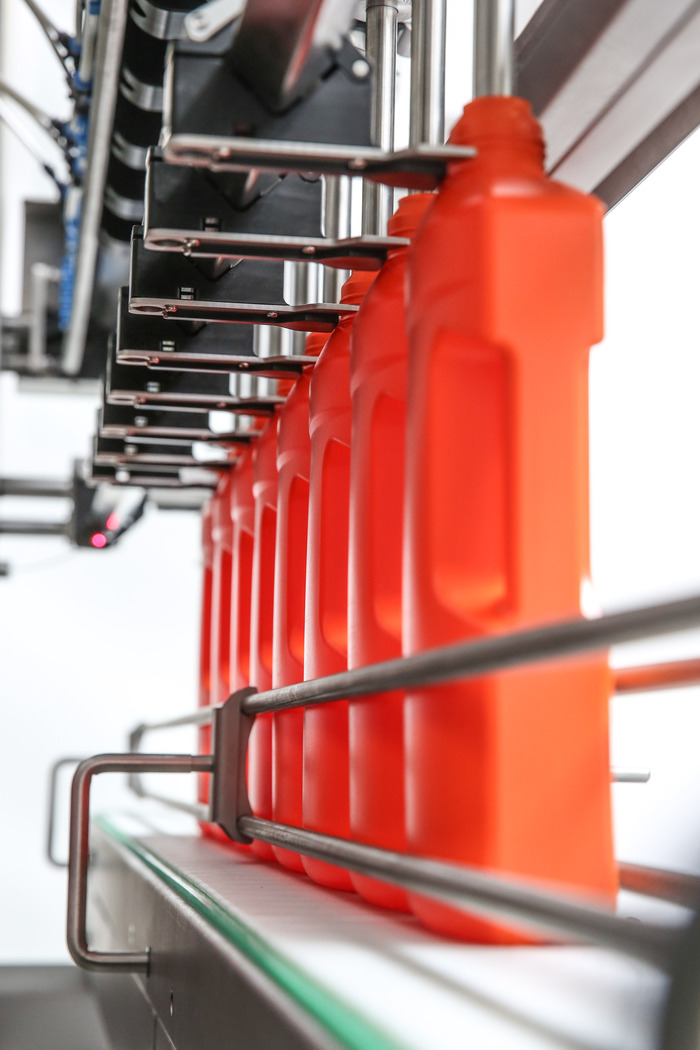 A row of orange plastic bottles is being conveyed on an automated conveyor belt in a manufacturing facility, undergoing the filing process.