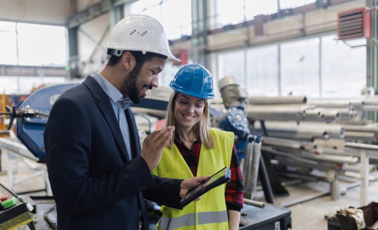 Two people in a factory setting, wearing safety helmets and safety gear, are looking at an Obeeco tablet and smiling. Pipes and industrial equipment can be seen in the background.