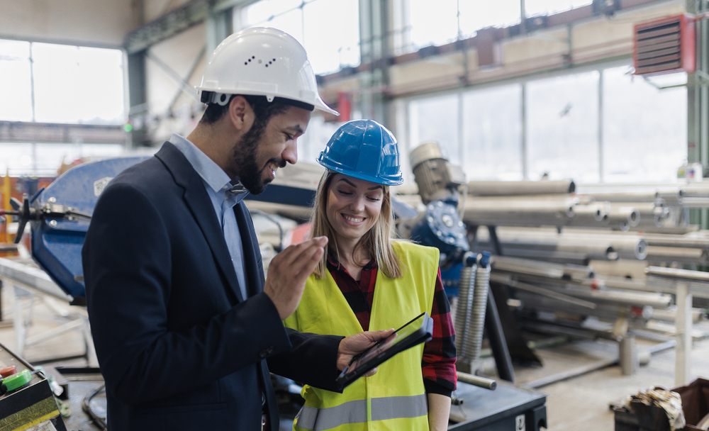 Two people in a factory setting, wearing safety helmets and safety gear, are looking at an Obeeco tablet and smiling. Pipes and industrial equipment can be seen in the background.