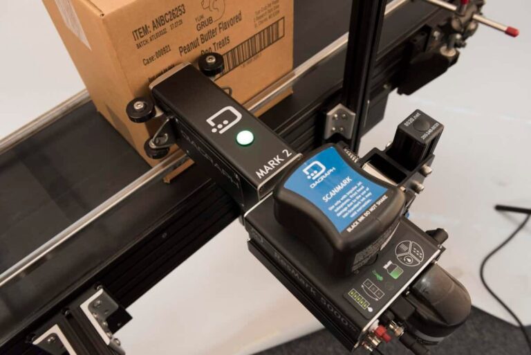 A conveyor belt with a box labeled "Peanut Butter Flavored Dog Treats" glides past the MARK-2 scanner, efficiently coding and marking each package in the industrial setting.